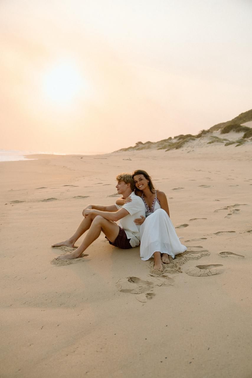 Couple sitting together on sandy beach dunes watching the golden sunset in peaceful embrace.