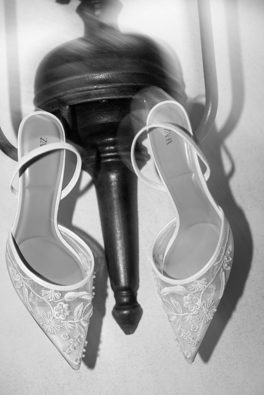 Black and white overhead view of delicate lace bridal heels with floral embroidery resting on dark wooden chair.