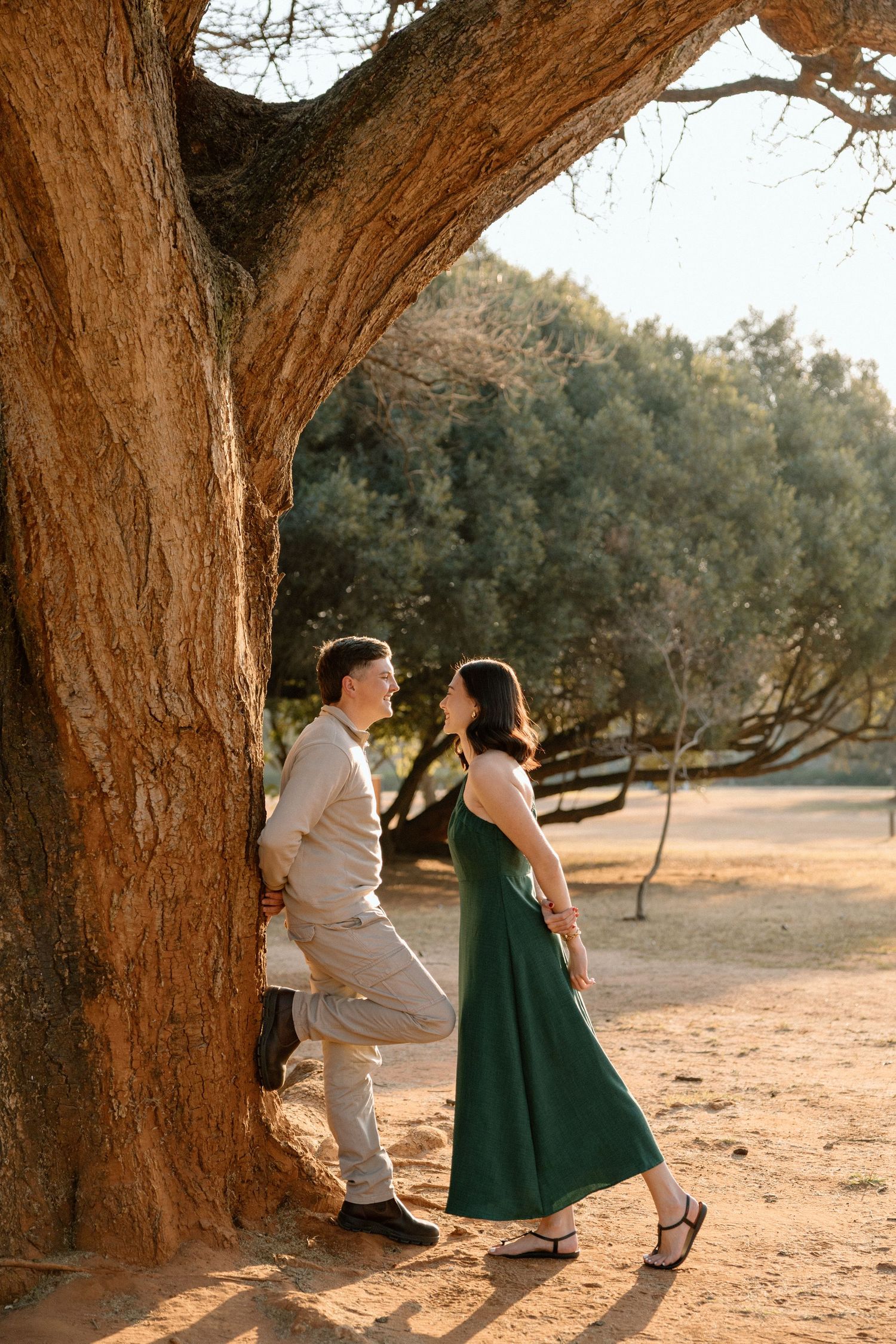 Couple in casual attire sharing a romantic moment leaning against a large tree at golden hour.