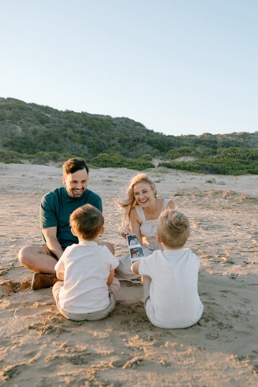 Happy family with two young children sitting on beach sand playing with a polaroid camera.