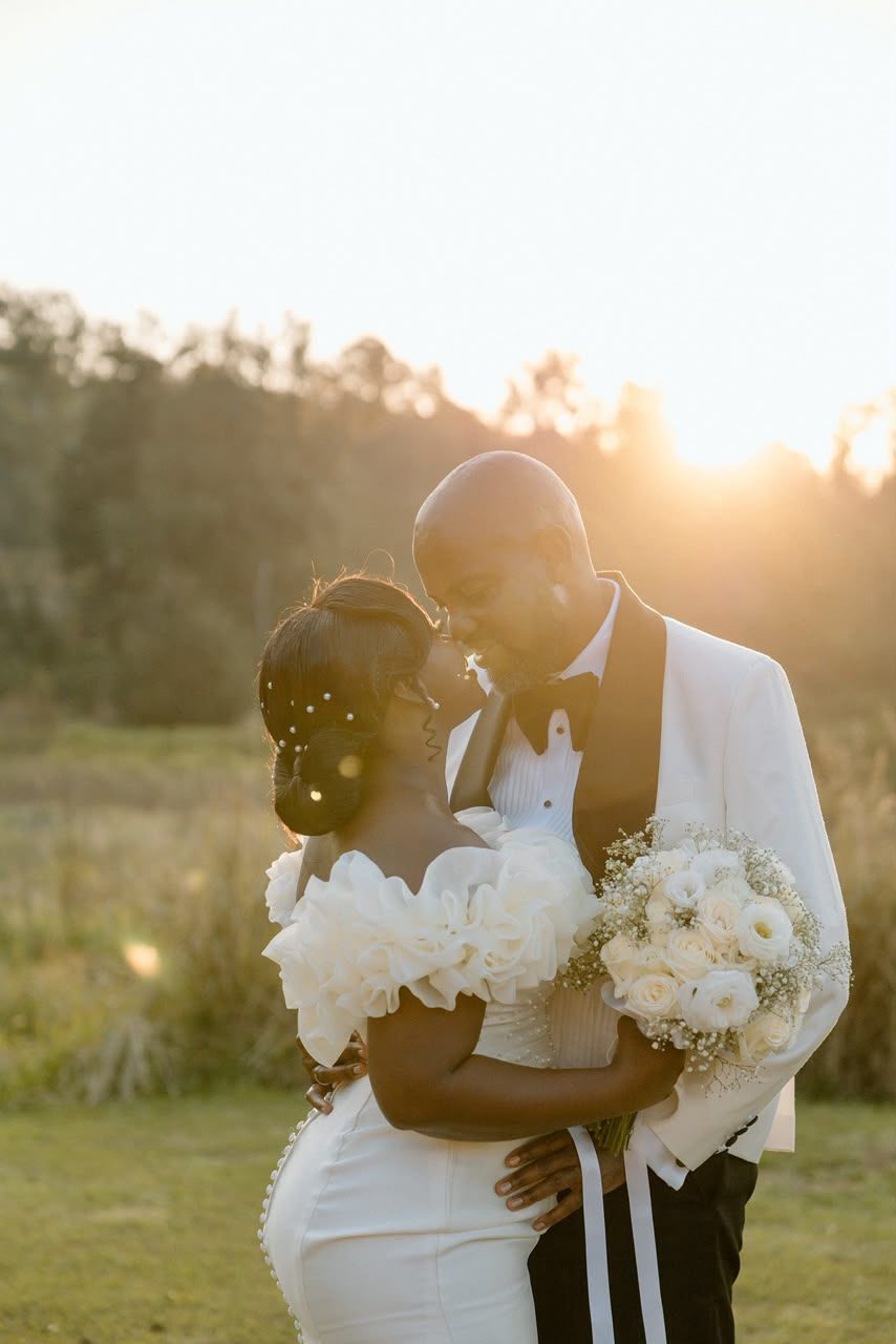 Bride and groom share romantic golden hour embrace with white bouquet in outdoor setting.
