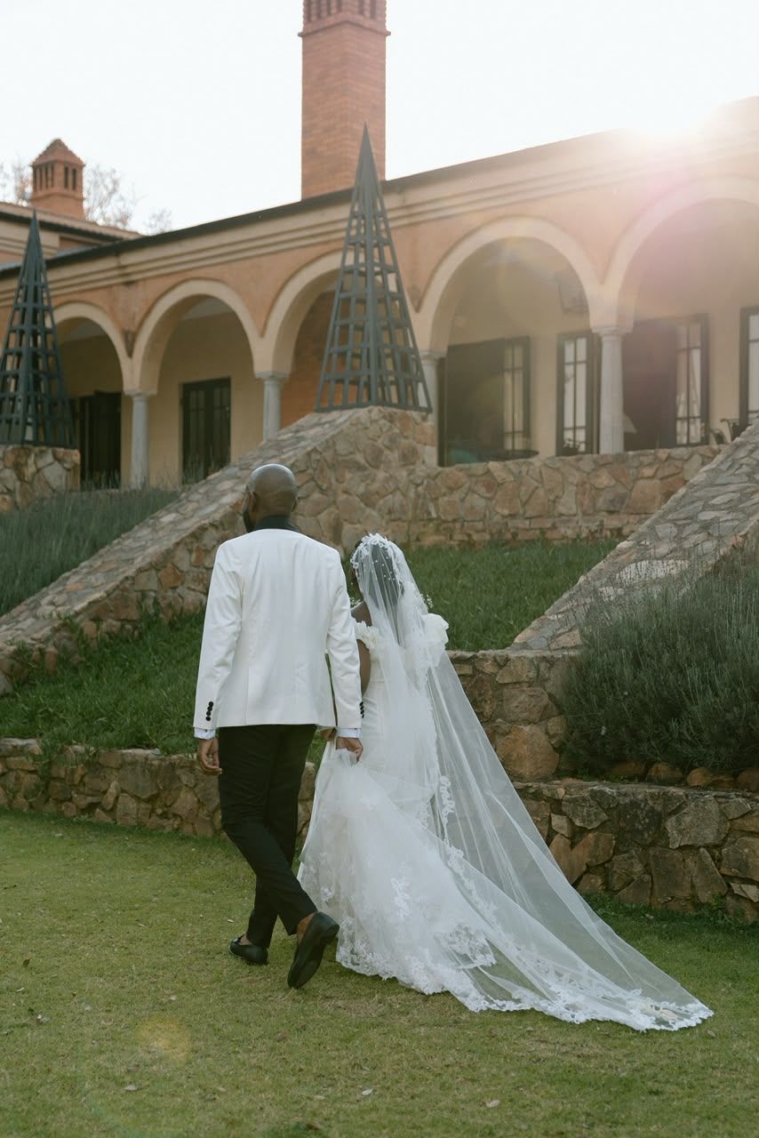 Bride and groom walking hand in hand toward elegant stone villa with arched windows in golden sunlight.