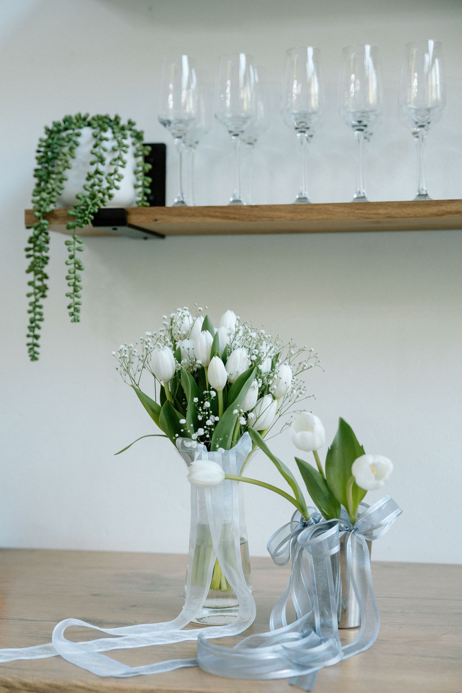 White tulip bridal bouquets with silver ribbon beside champagne flutes on wooden shelf display.