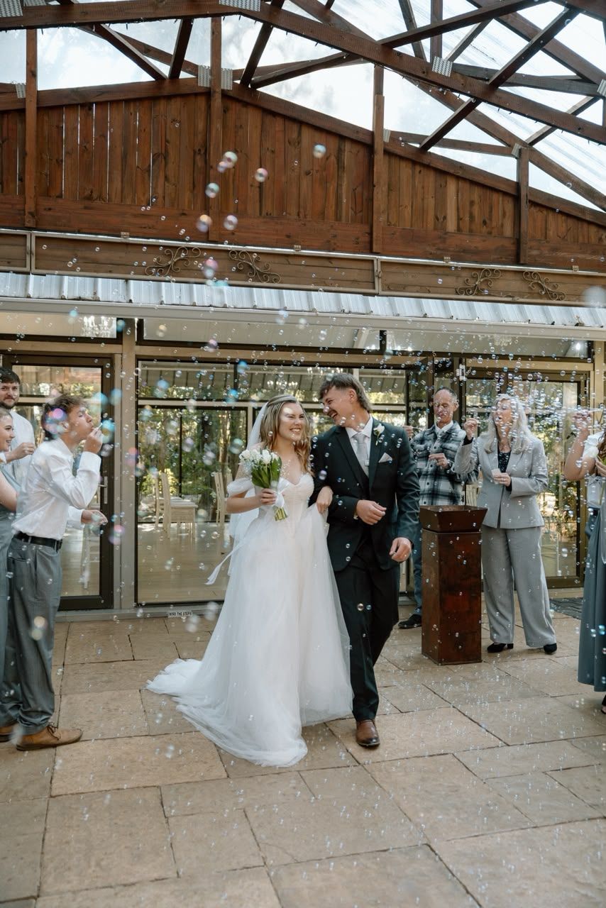 Newlyweds walk through joyful bubble send-off surrounded by cheering guests outside wedding venue.