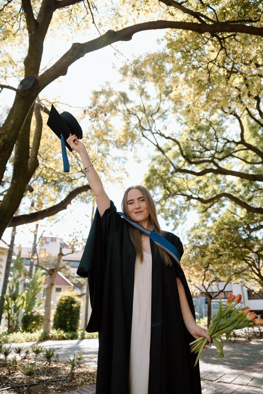 Happy female graduate in black gown raises cap triumphantly while holding tulips under trees.