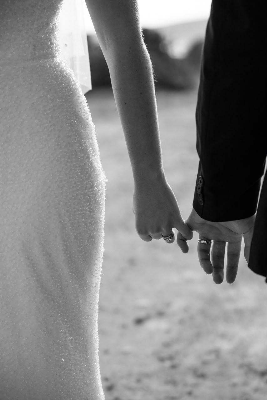 Intimate black and white close-up of bride and groom holding hands showing wedding rings during outdoor ceremony.