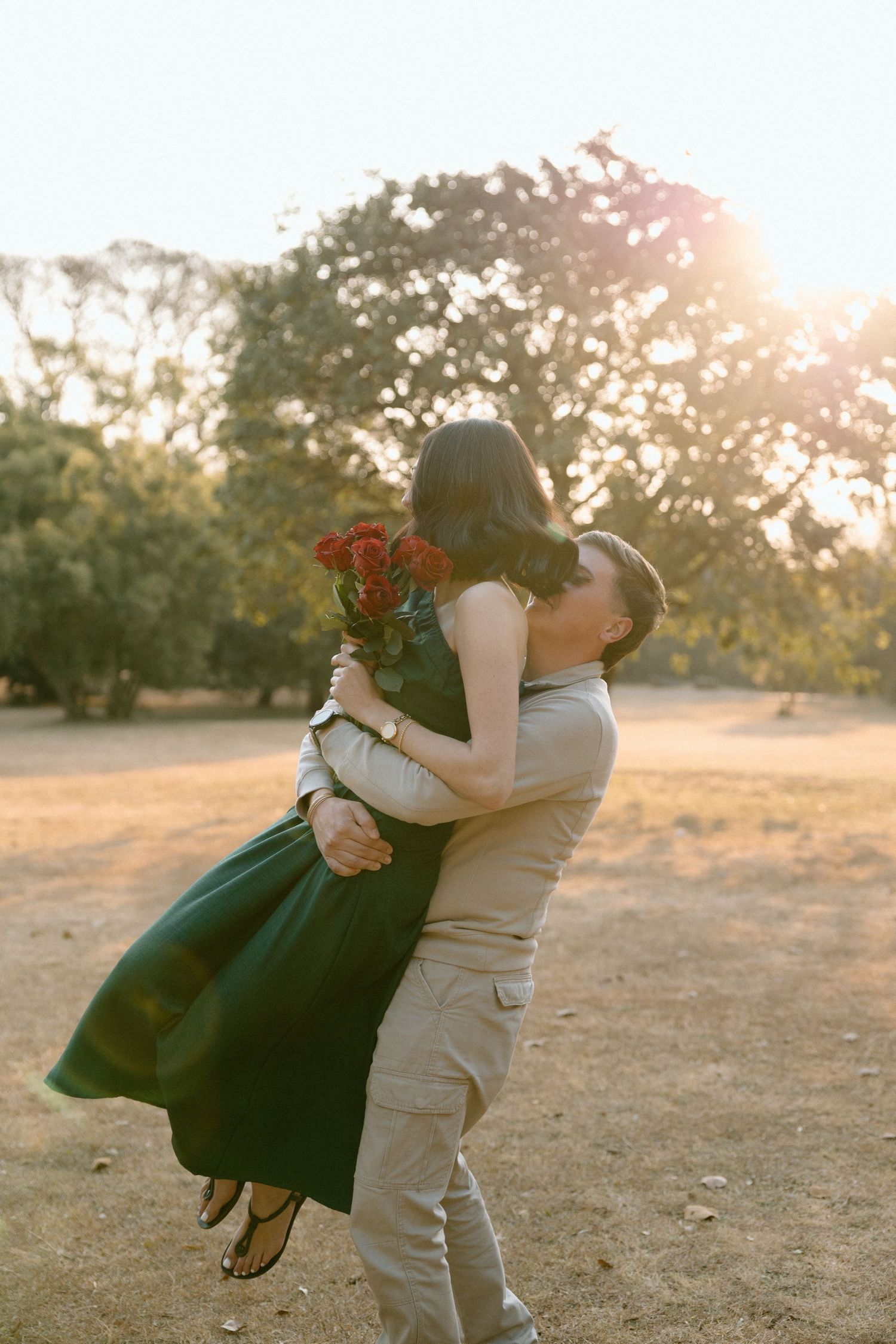 Man lifting woman in green dress holding red roses bouquet, sharing a kiss in golden sunset light.