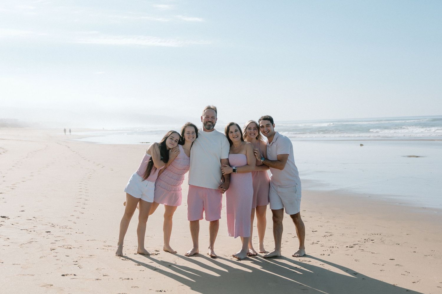 Happy family of six poses together smiling on sunny ocean beach in coordinated outfits.