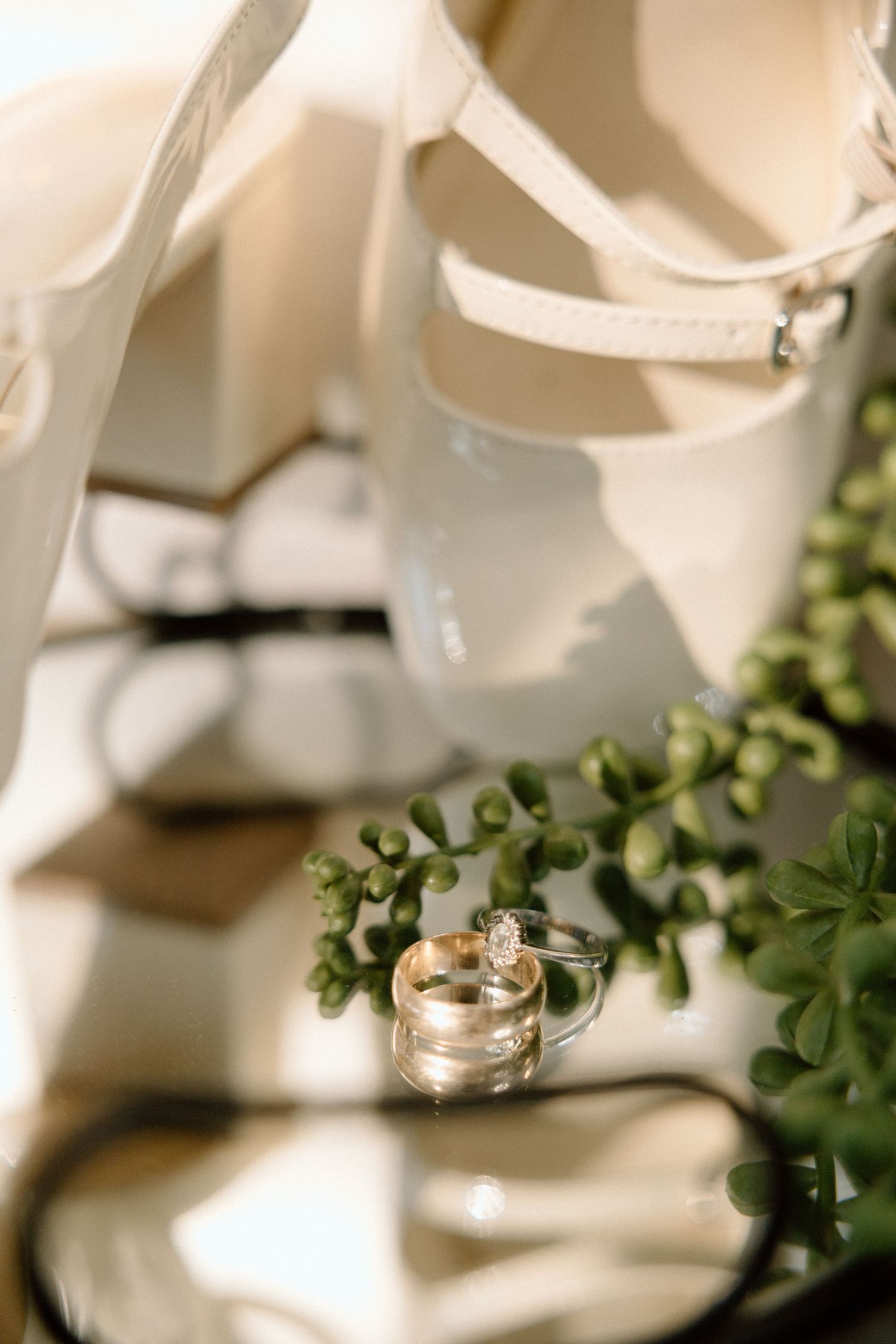 Close-up of gold wedding bands and diamond engagement ring beside white bridal heels and greenery.