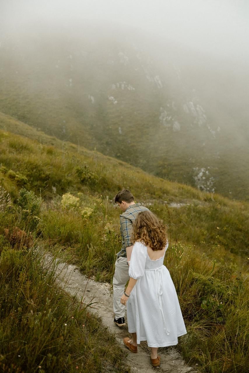 Couple holding hands while hiking a narrow stone path on a foggy green mountain hillside.