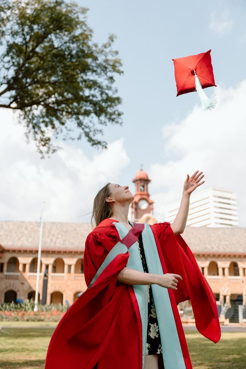 Graduate in red doctoral gown throws red mortarboard cap into sky on university grounds.