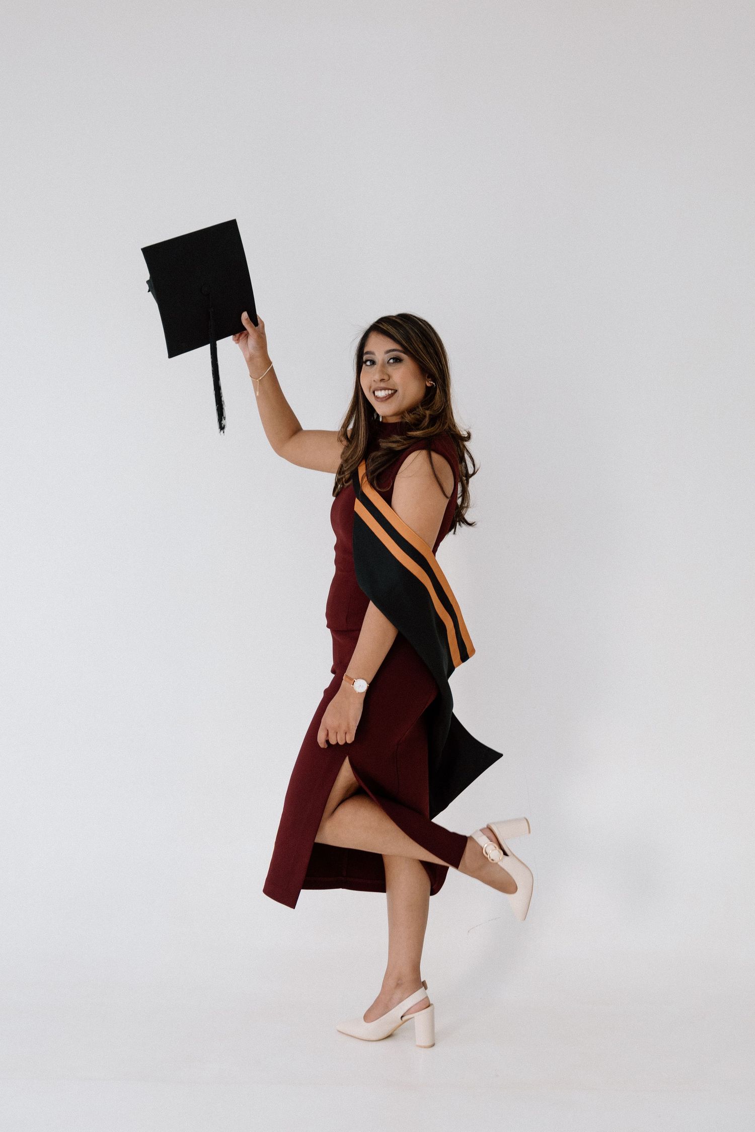 Smiling graduate in maroon dress holding cap aloft, wearing academic hood with gold stripe, celebrating graduation.