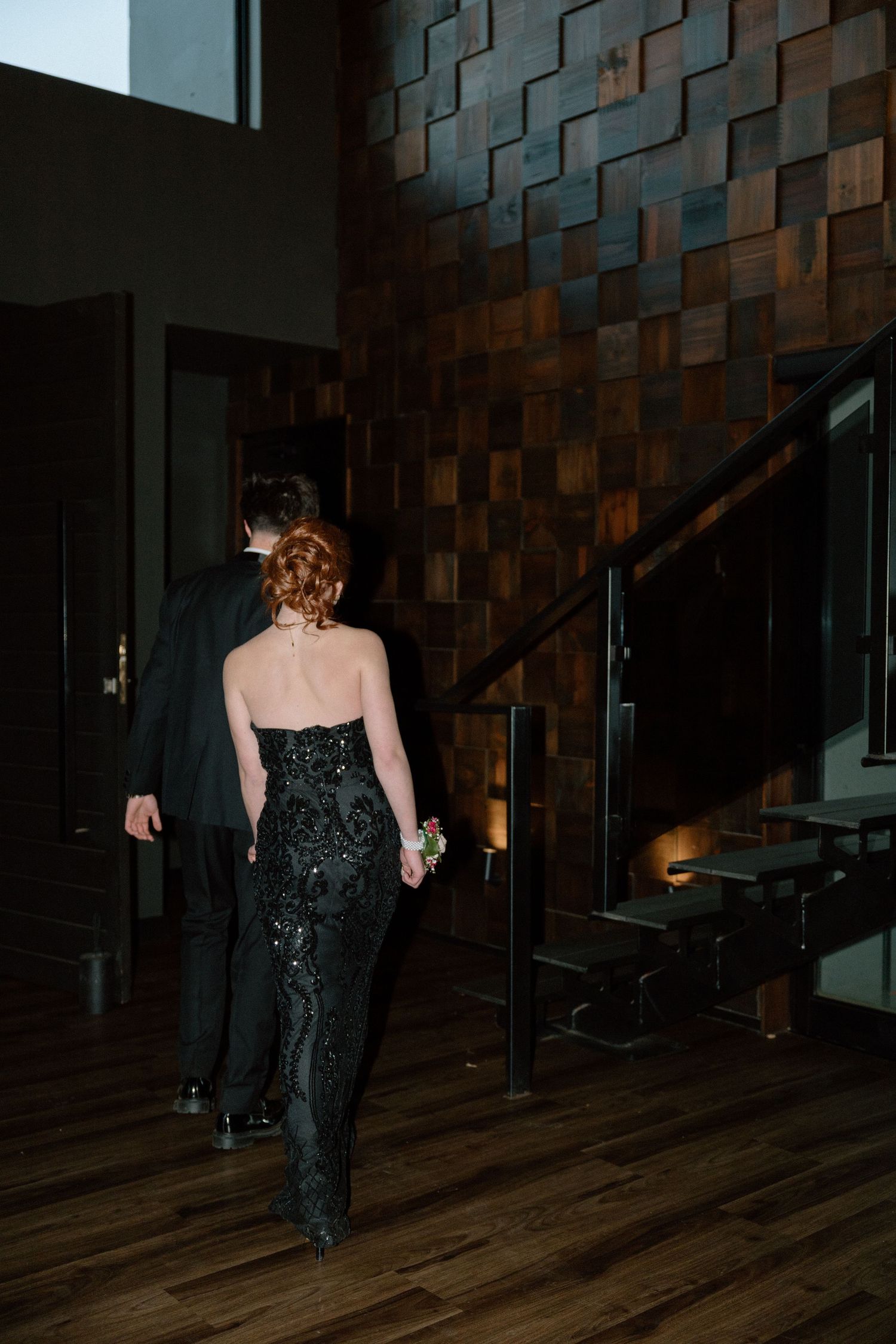 Couple in formal black attire walking through elegant venue with geometric wood wall and staircase.