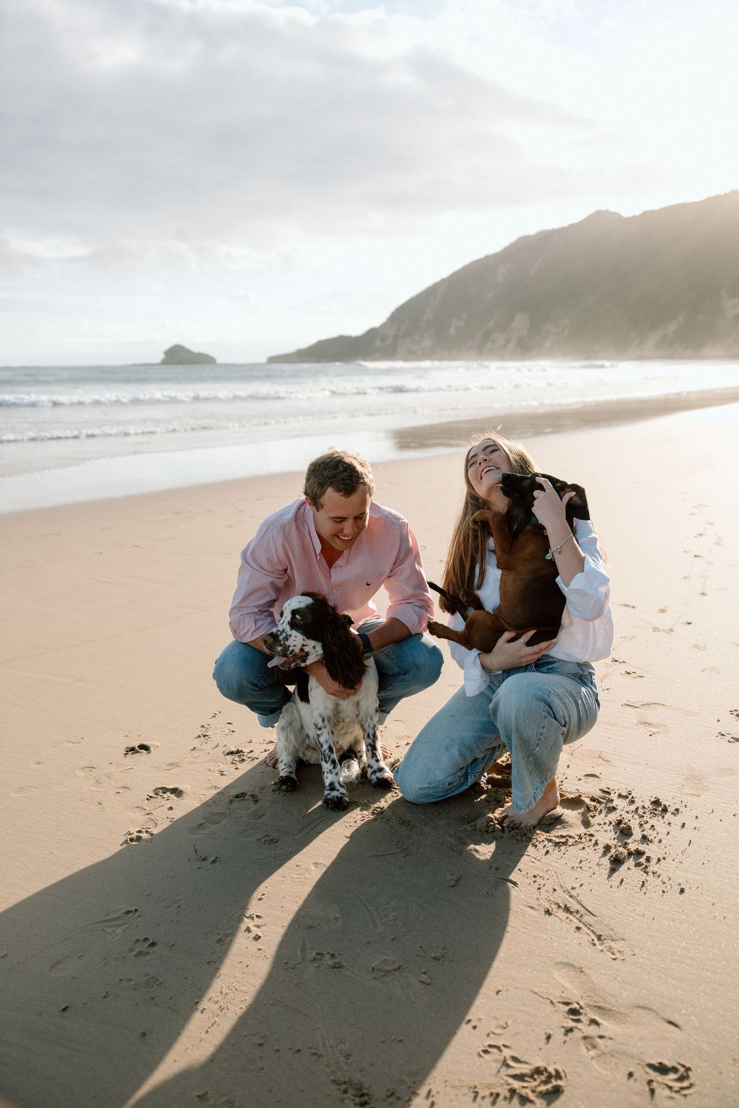 Couple crouching on sandy beach with their Springer Spaniel dog at golden hour near coastal cliffs.