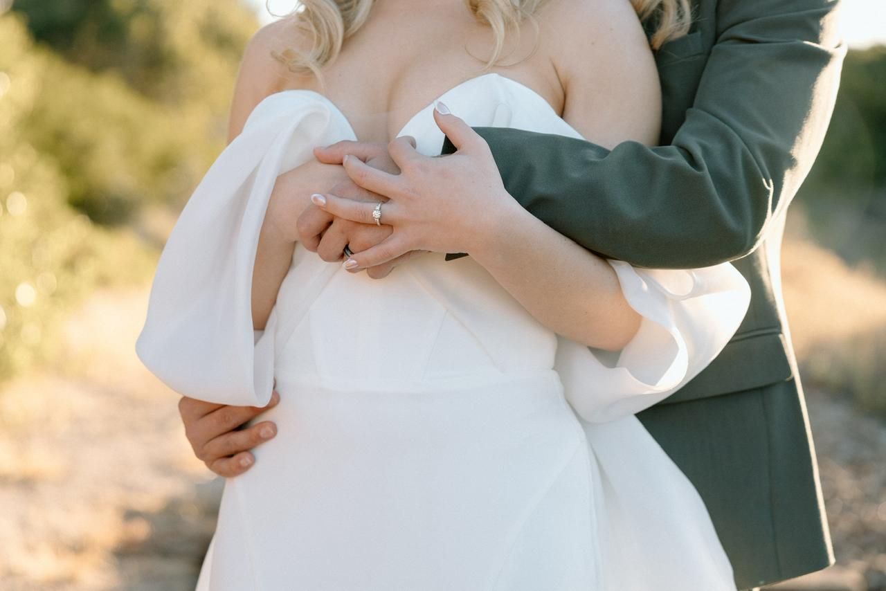 Close-up of groom in grey suit embracing bride in white off-shoulder dress showing wedding ring.