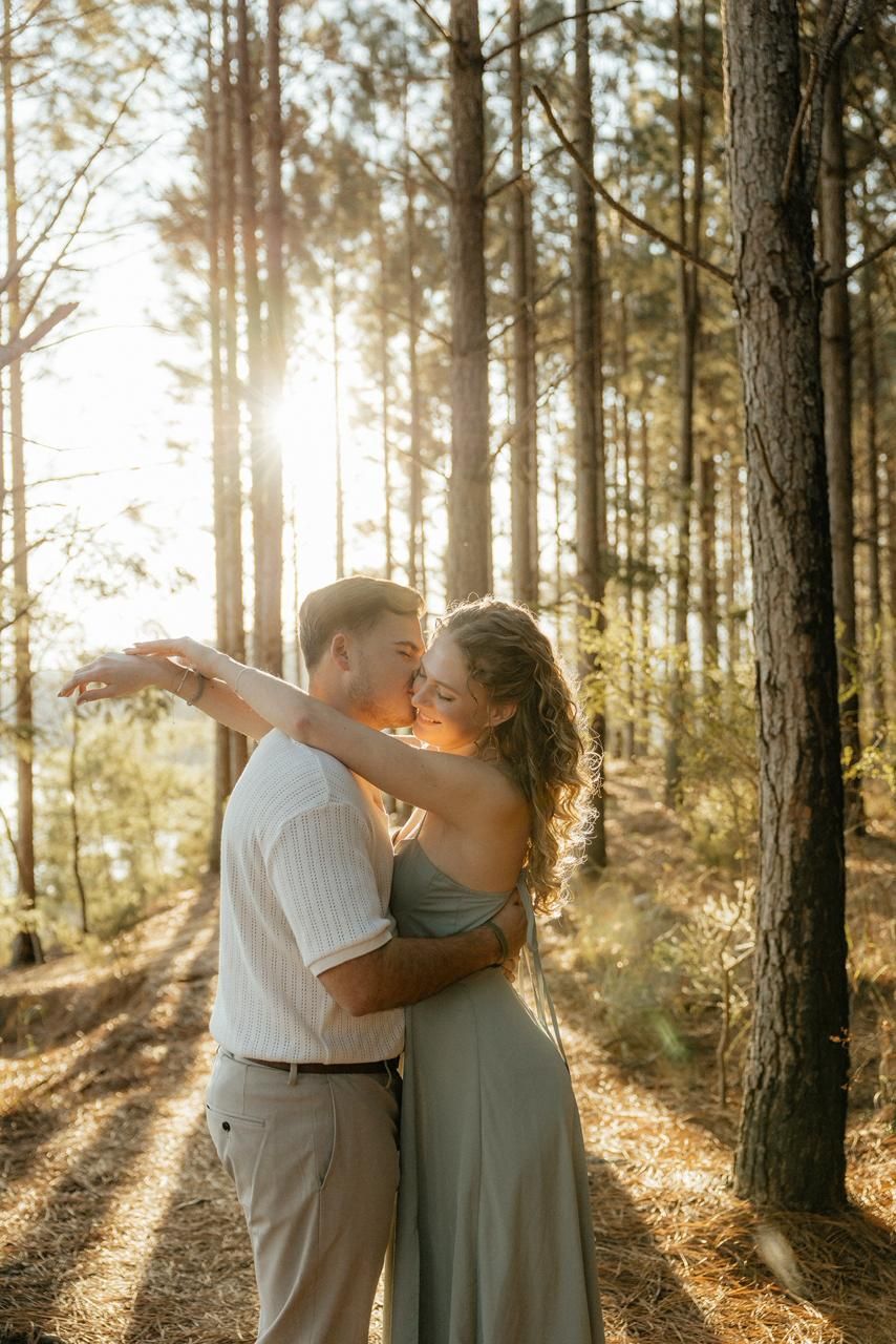 Couple sharing a kiss while embracing in a sun-drenched pine forest with golden backlight.