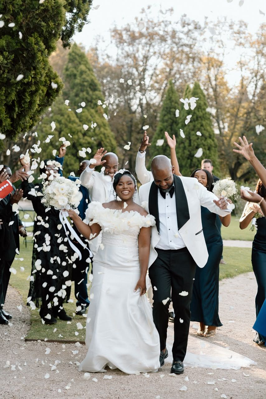 Joyful newlywed couple celebrating with wedding party tossing white flower petals in lush garden setting.