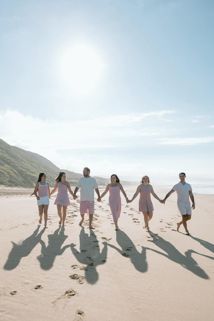 Group of six friends holding hands walking together along sunny beach with coastal hills behind.
