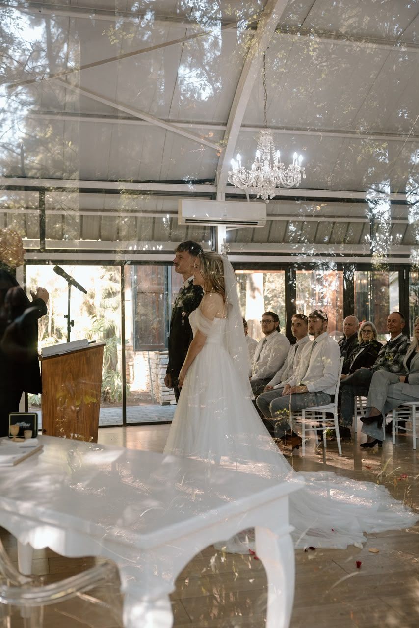 Bride and groom share a kiss at the altar during an elegant indoor wedding ceremony under a glass ceiling.
