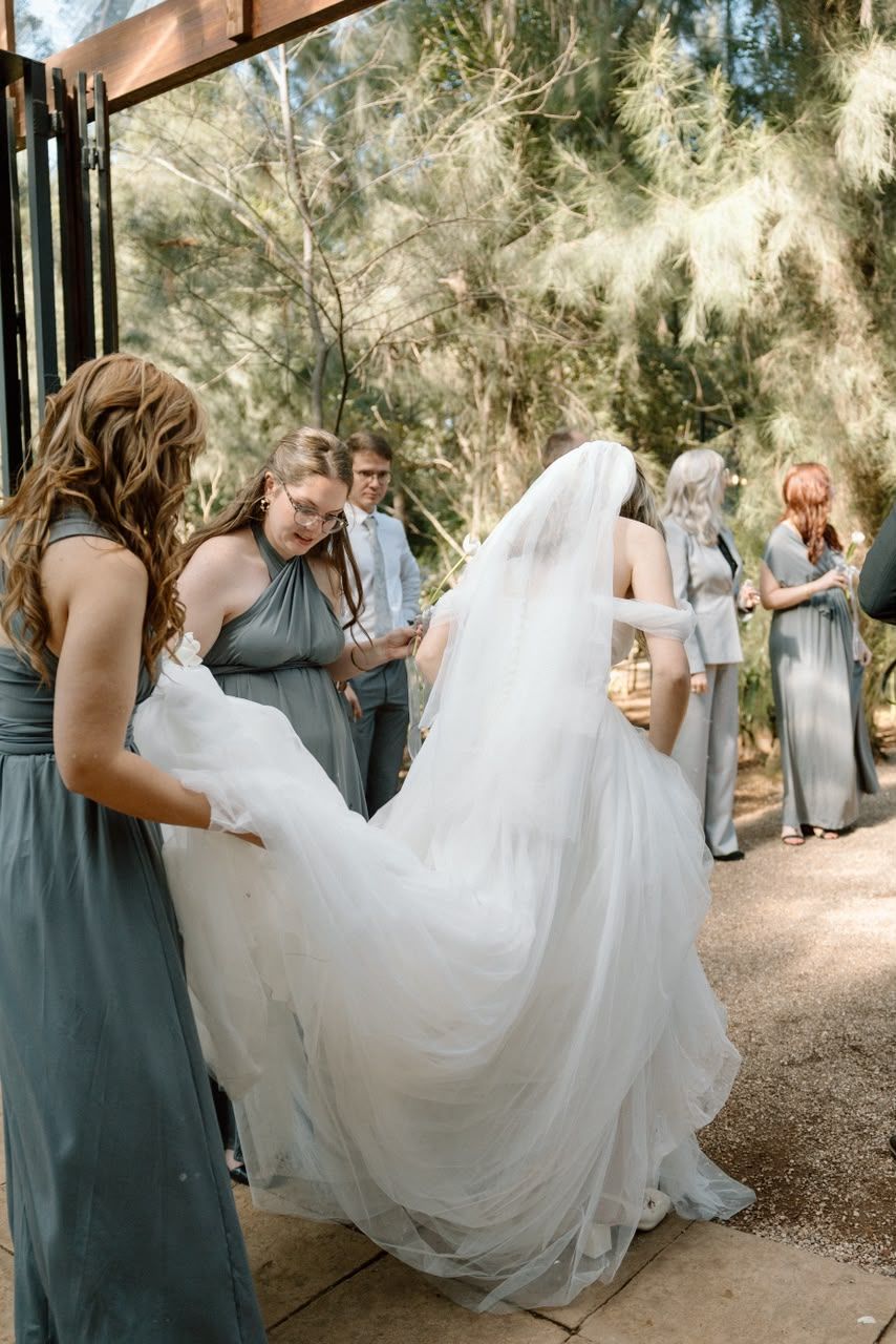 Bridesmaids in sage dresses help arrange brides flowing white veil and train before outdoor ceremony.