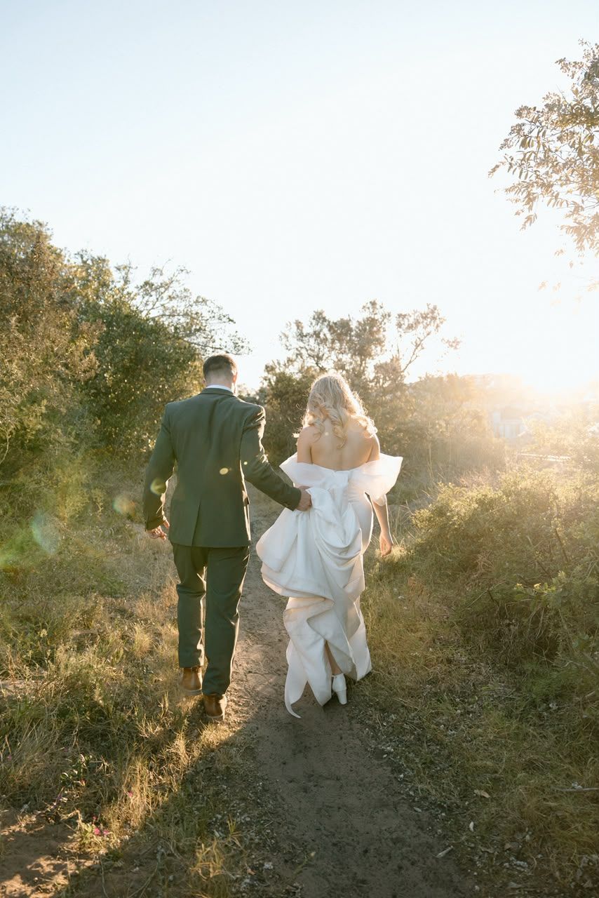 A bride and groom walk hand-in-hand down a sunlit path surrounded by golden foliage on their wedding day.