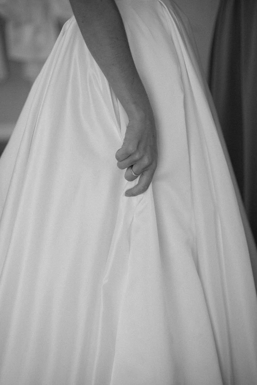 Black and white photo of a bride holding her flowing white wedding gown, with a ring visible on her finger.