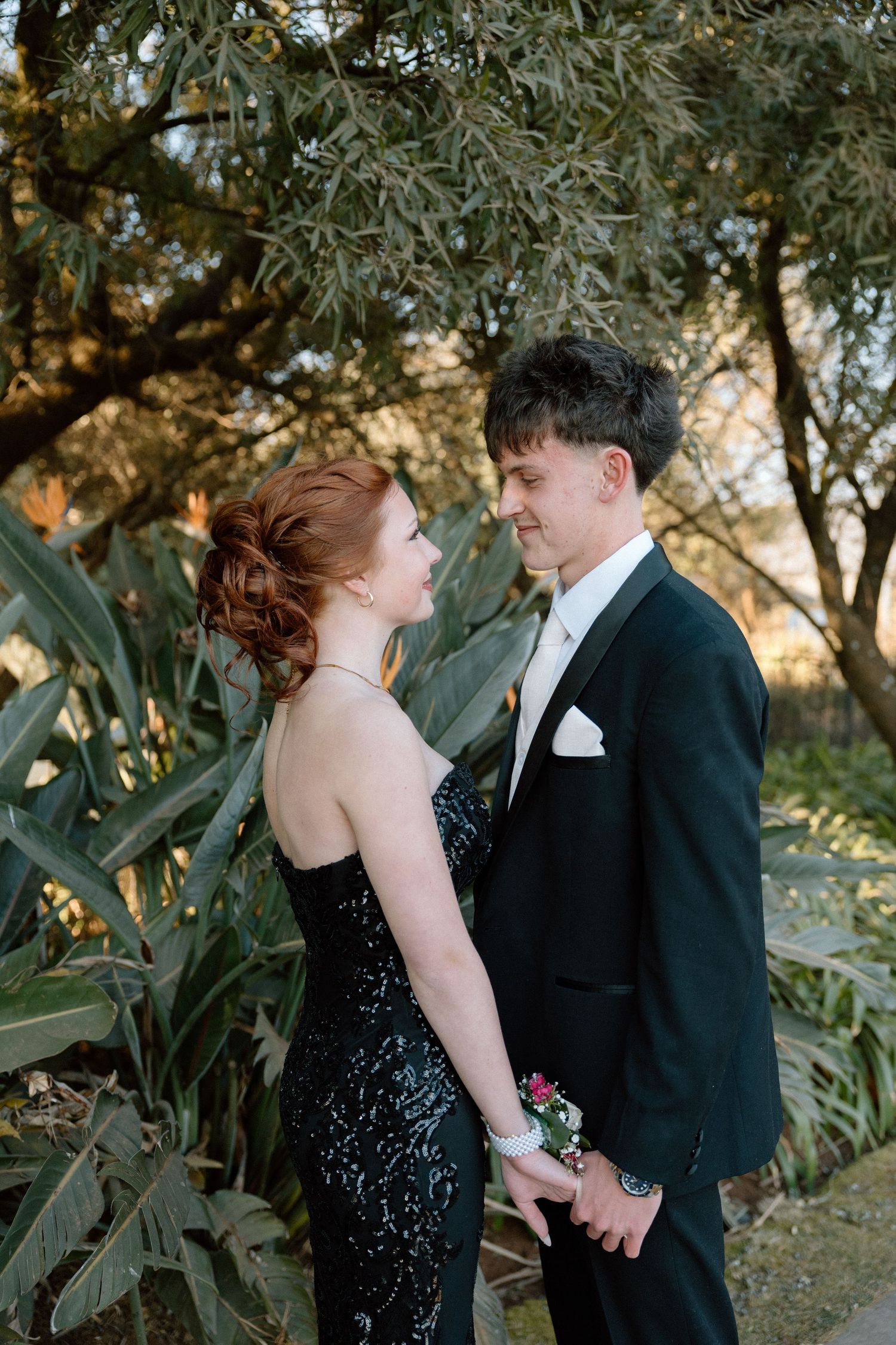A young couple in formal attire holds hands outdoors, she in a black gown and he in a tuxedo, posing for prom photos.