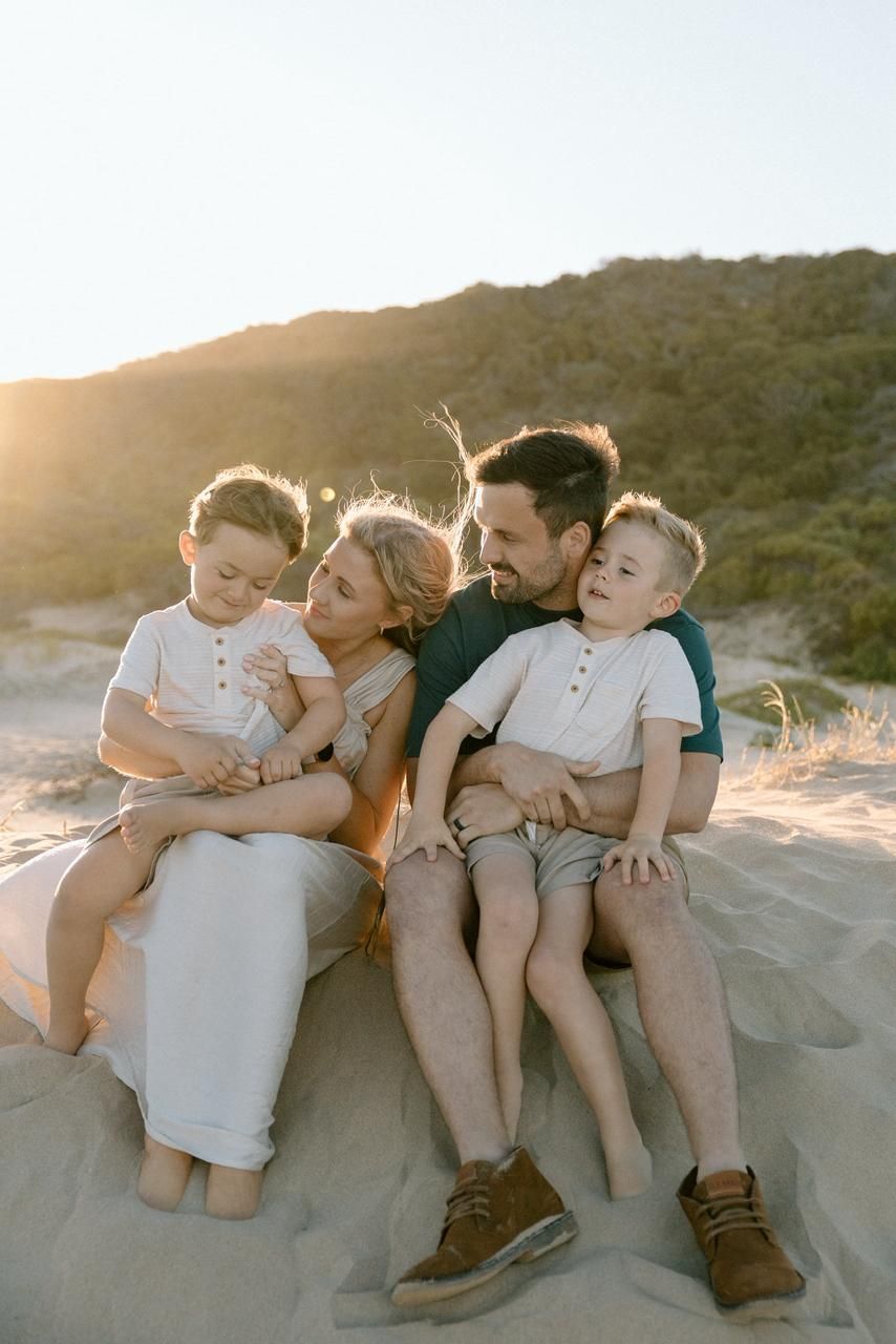 Family of four with two young boys sitting together on sandy beach at golden sunset.