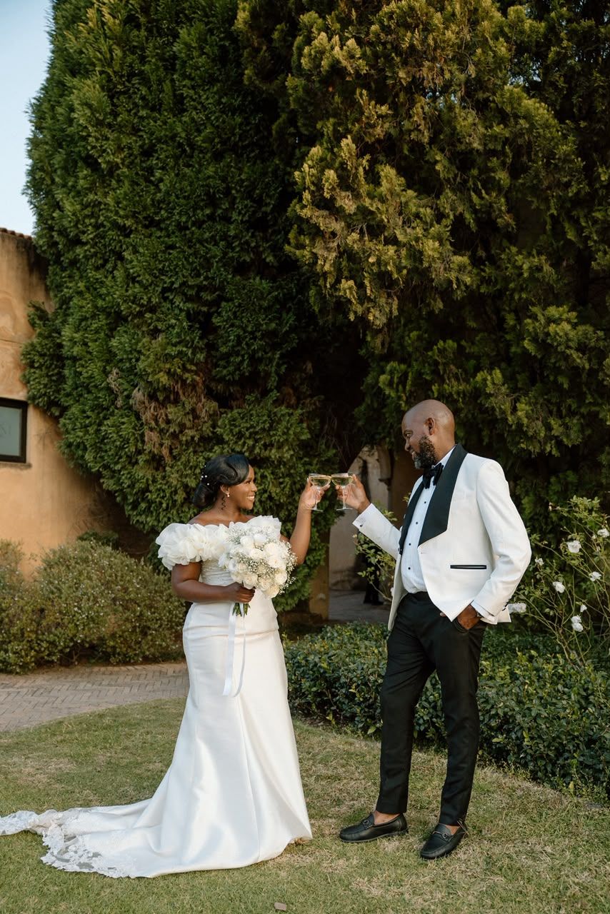 Elegant Black couple toasting champagne in garden, bride in off-shoulder gown, groom in white tuxedo jacket.