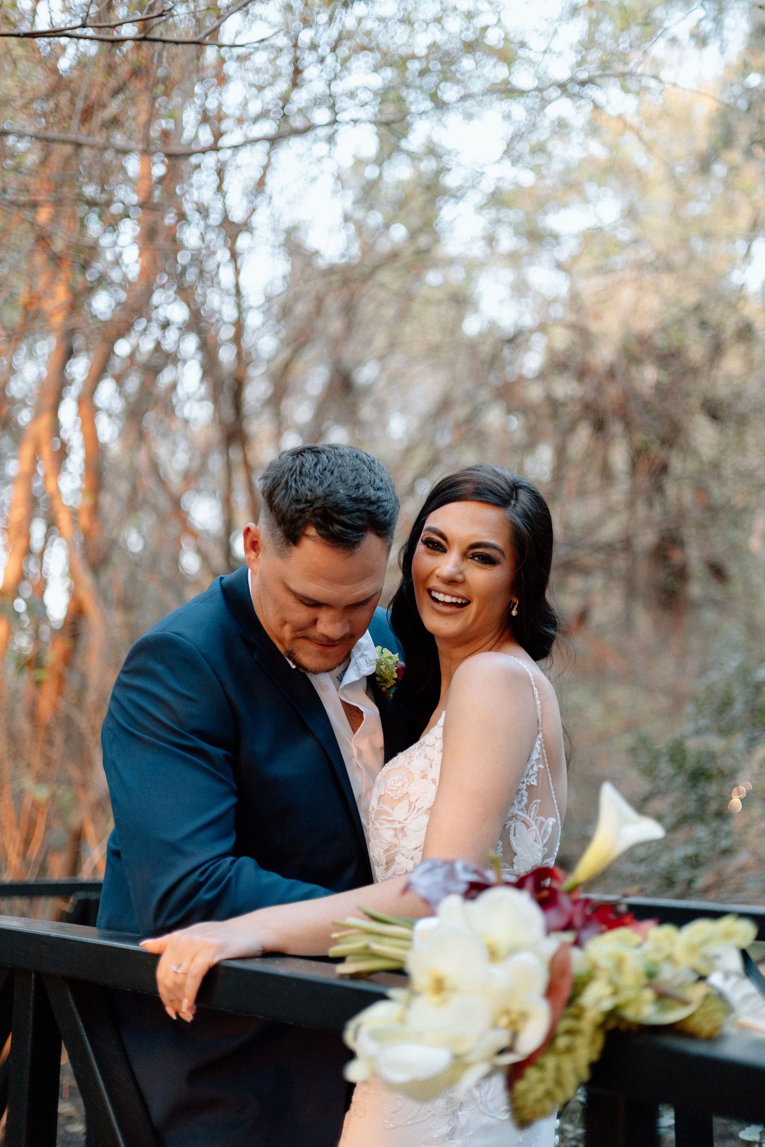 A bride and groom share a joyful moment on a wooden bridge surrounded by autumn trees, bride holding a white floral bouquet.