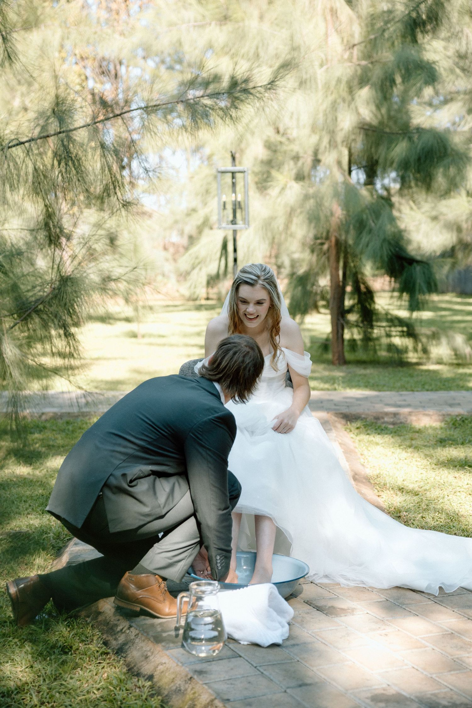 Groom washing bride's feet in outdoor wedding ceremony surrounded by pine trees.