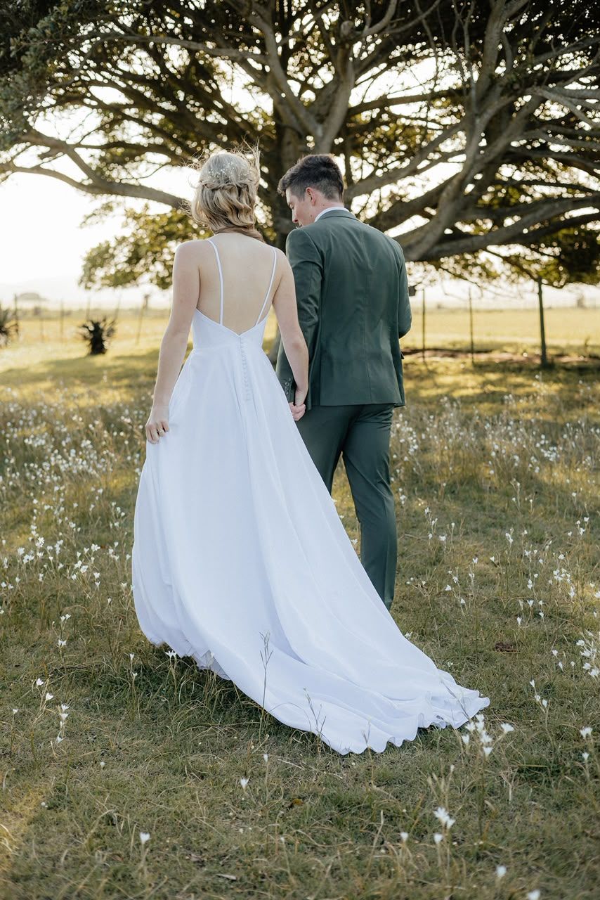 Bride in white gown and groom in green suit walk hand in hand through a sunlit field toward a large tree.
