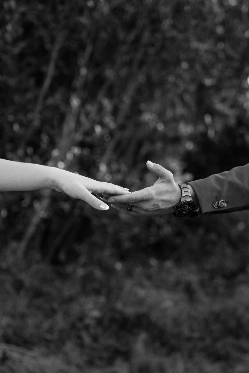 Black and white photo of two hands almost touching in a romantic gesture, with blurred foliage in the background.