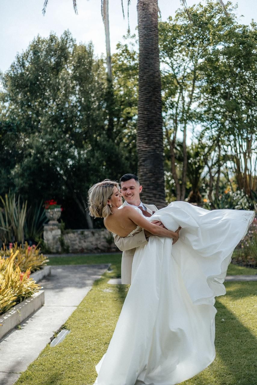 Groom lifts smiling bride in white wedding gown in lush tropical garden with palm trees.