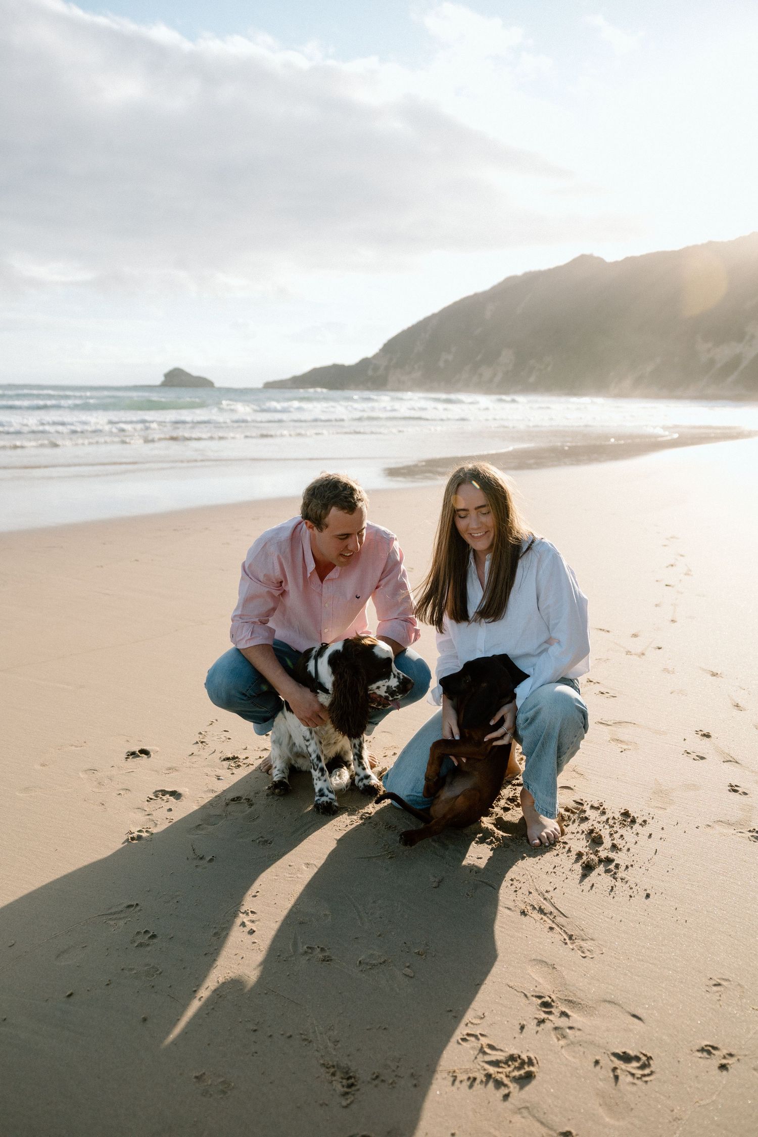 Smiling couple crouching on sandy beach petting two dogs with ocean waves and mountains in background.
