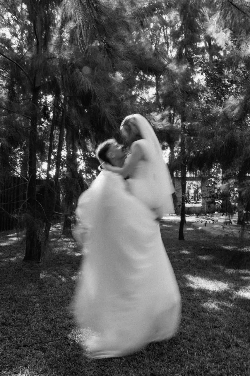 Black and white motion blur photo of bride and groom sharing a romantic dance kiss among pine trees.