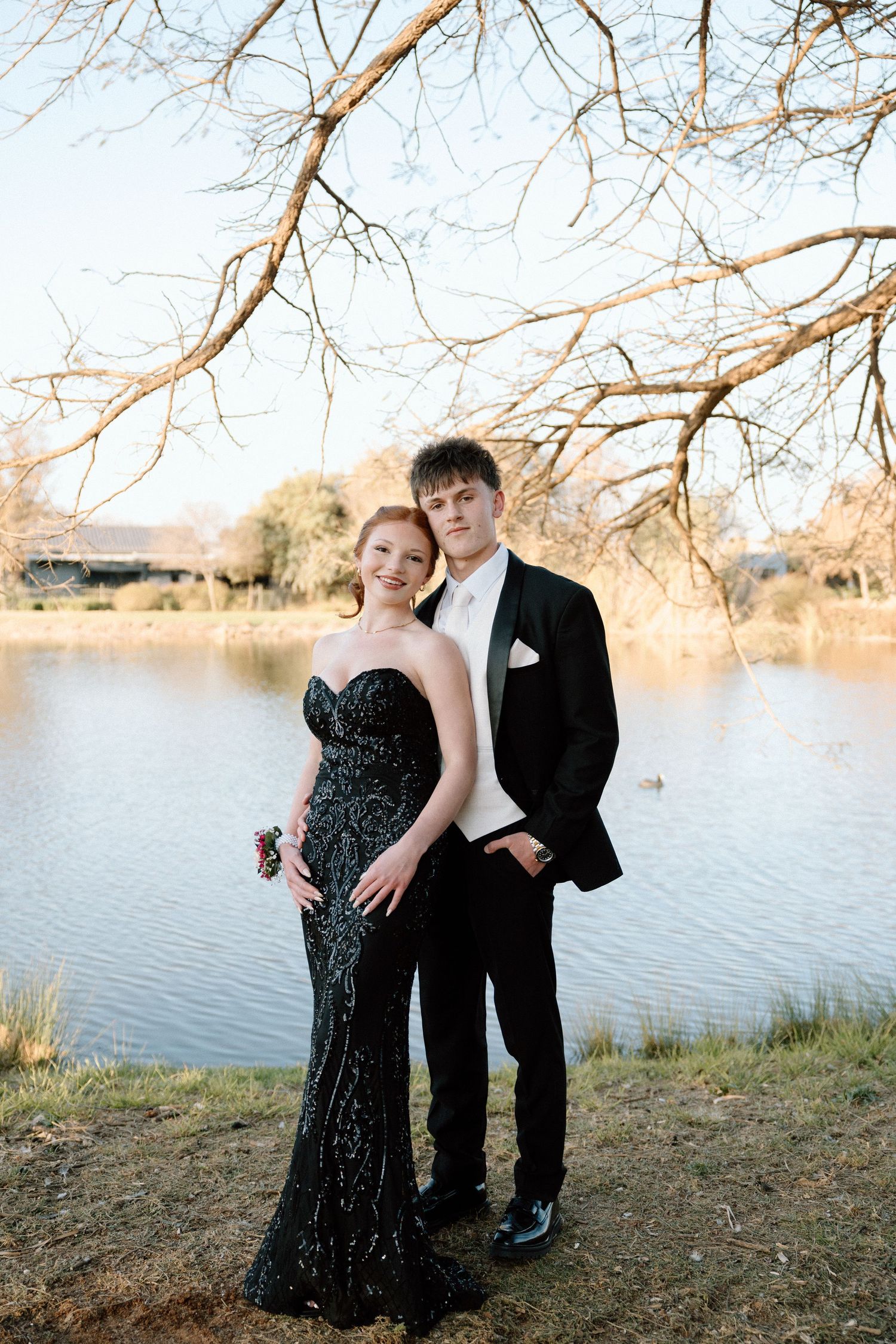 A young couple poses by a lake in formal prom attire, she in a black sequin gown and he in a classic tuxedo.