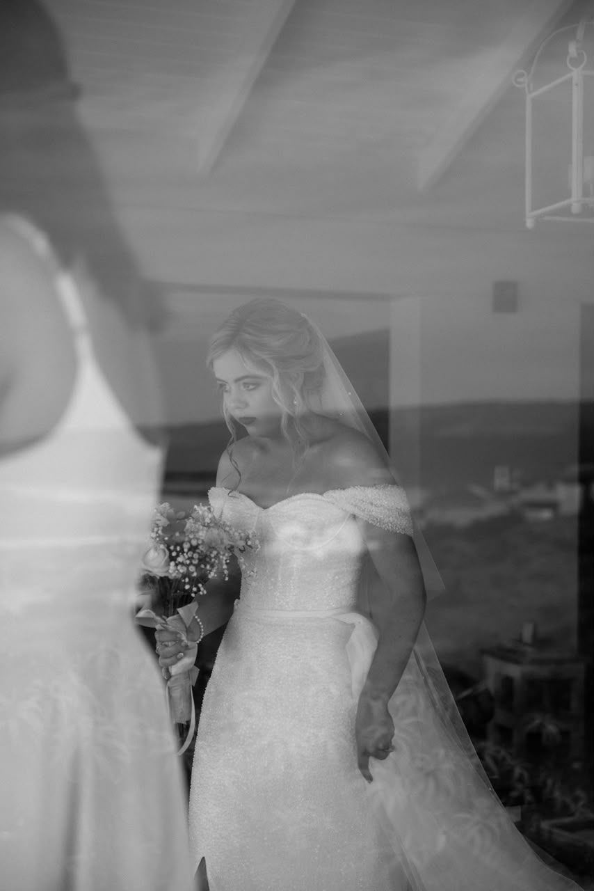 Elegant bride in off-shoulder lace wedding gown with veil holding bouquet, reflected in glass window.