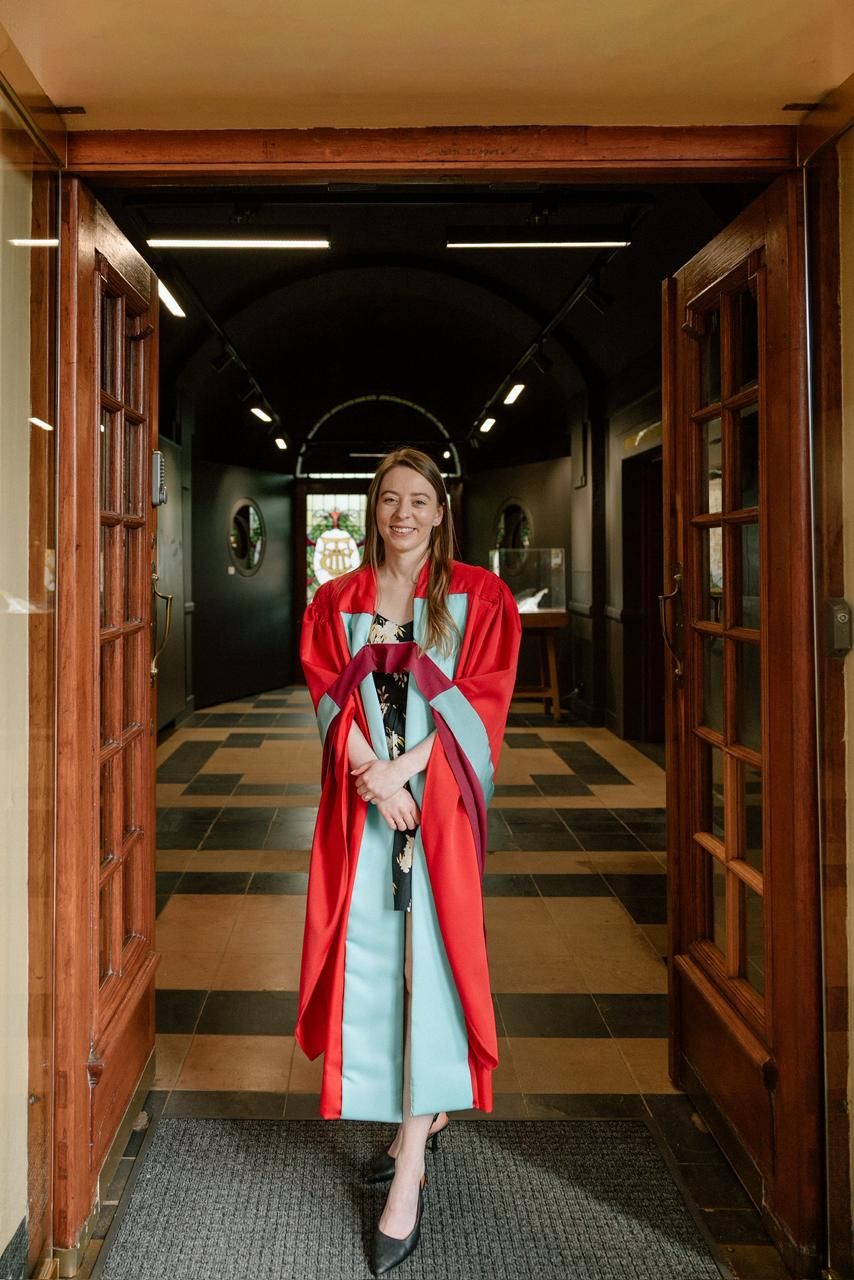 Smiling female graduate in red doctoral gown walks through elegant university corridor doorway.