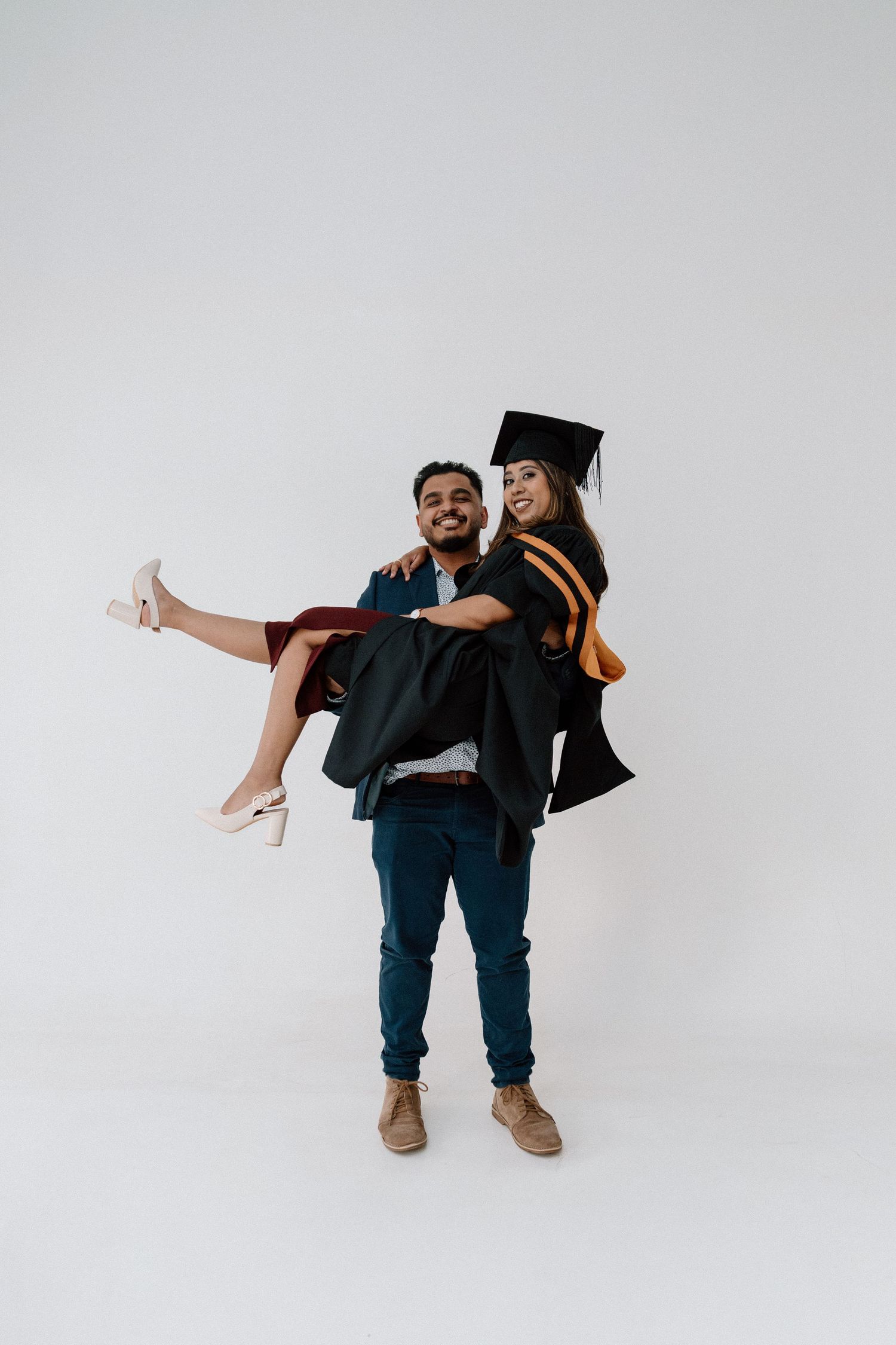 A smiling man carries a woman in graduation cap and gown in a celebratory pose against a white background.