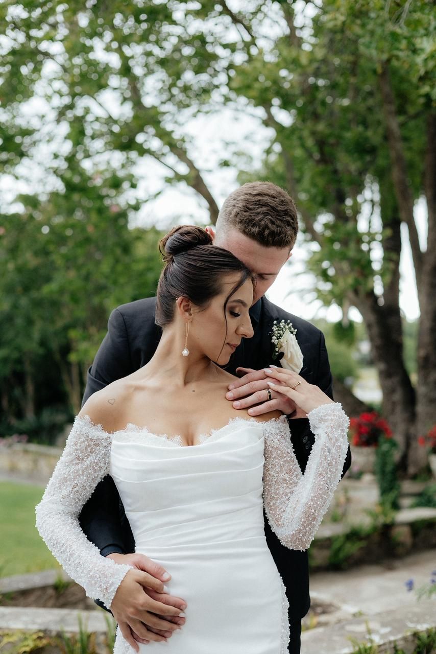 Groom tenderly embraces bride from behind in lace long-sleeve wedding dress among garden greenery.