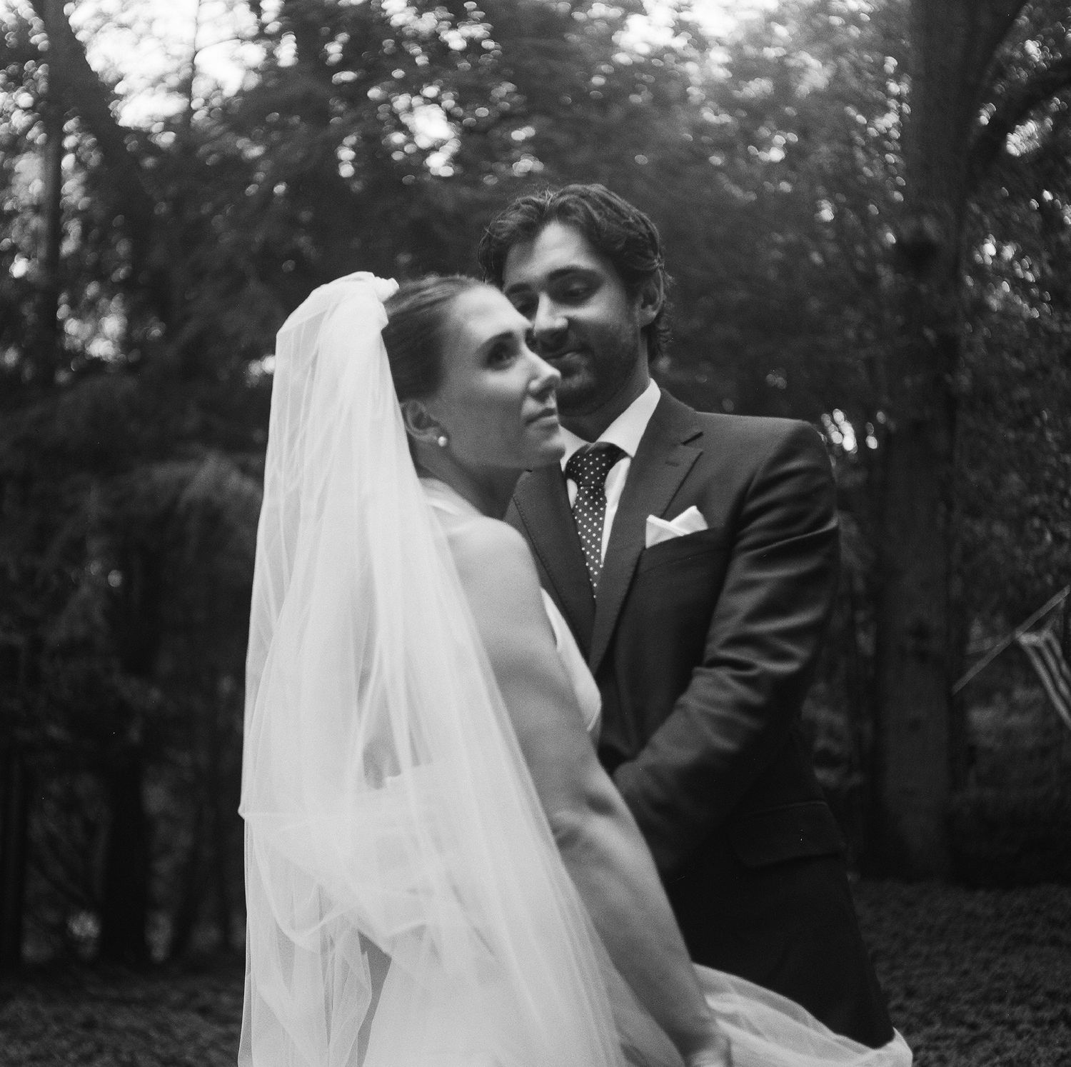 Black and white wedding photo of bride in veil and groom in suit embracing outdoors among trees.