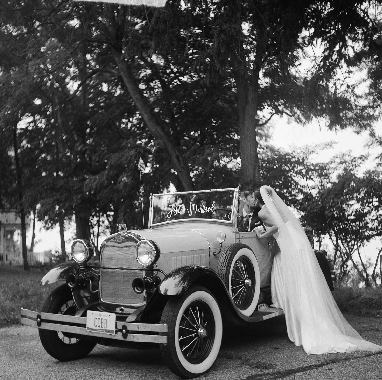 Vintage black and white photo of a bride in a white veil posing beside a classic antique convertible car outdoors.
