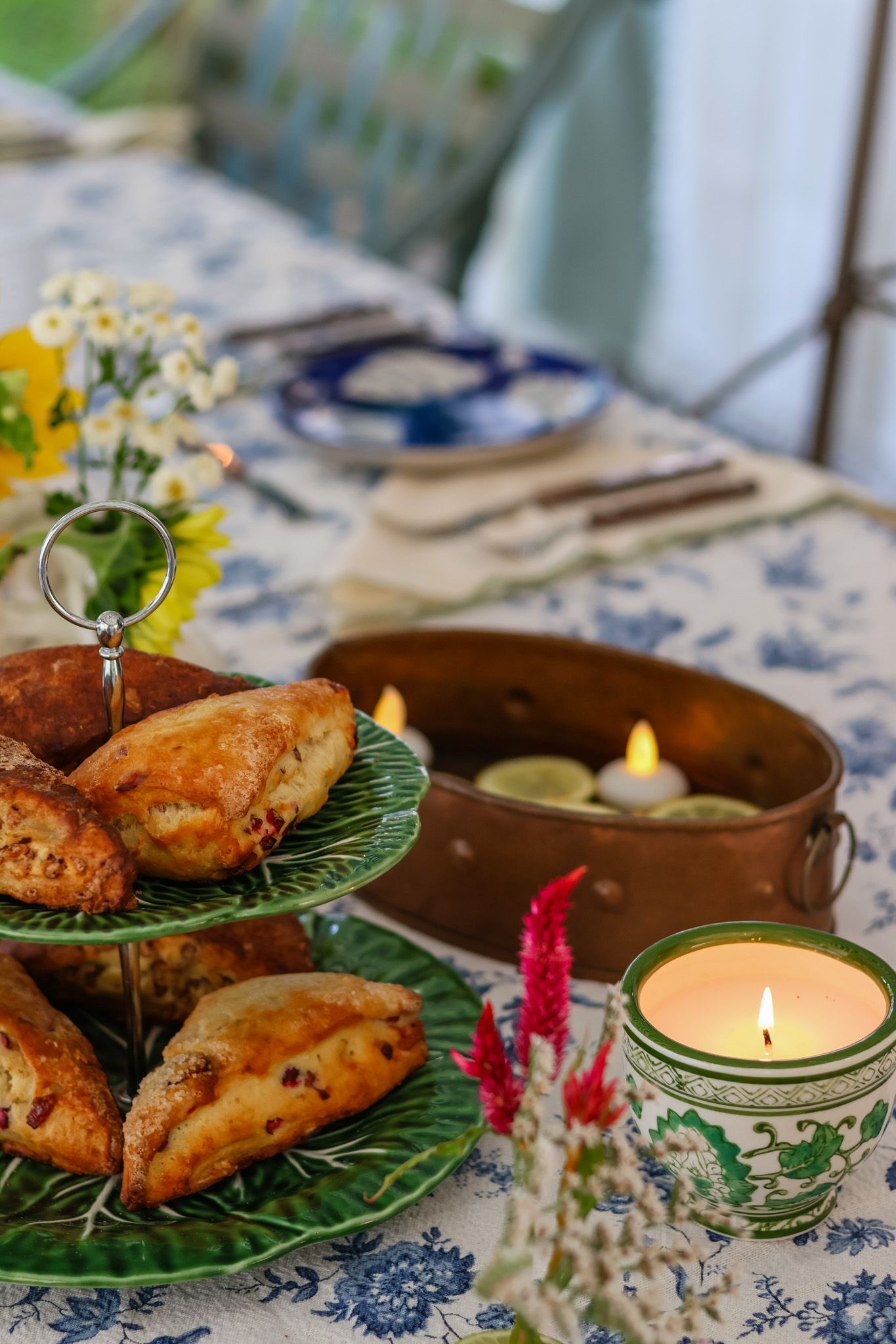 Tiered green serving stand with pastries beside lit candles on elegant blue floral tablecloth.
