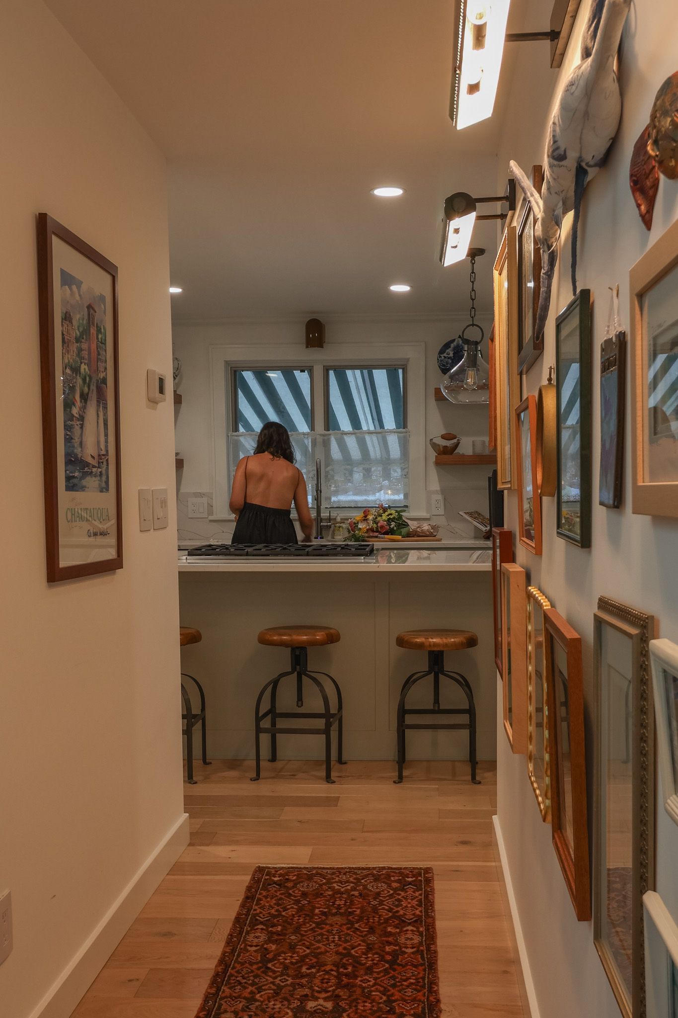 A woman cooking in a modern kitchen viewed through a hallway lined with framed artwork and warm wood floors.