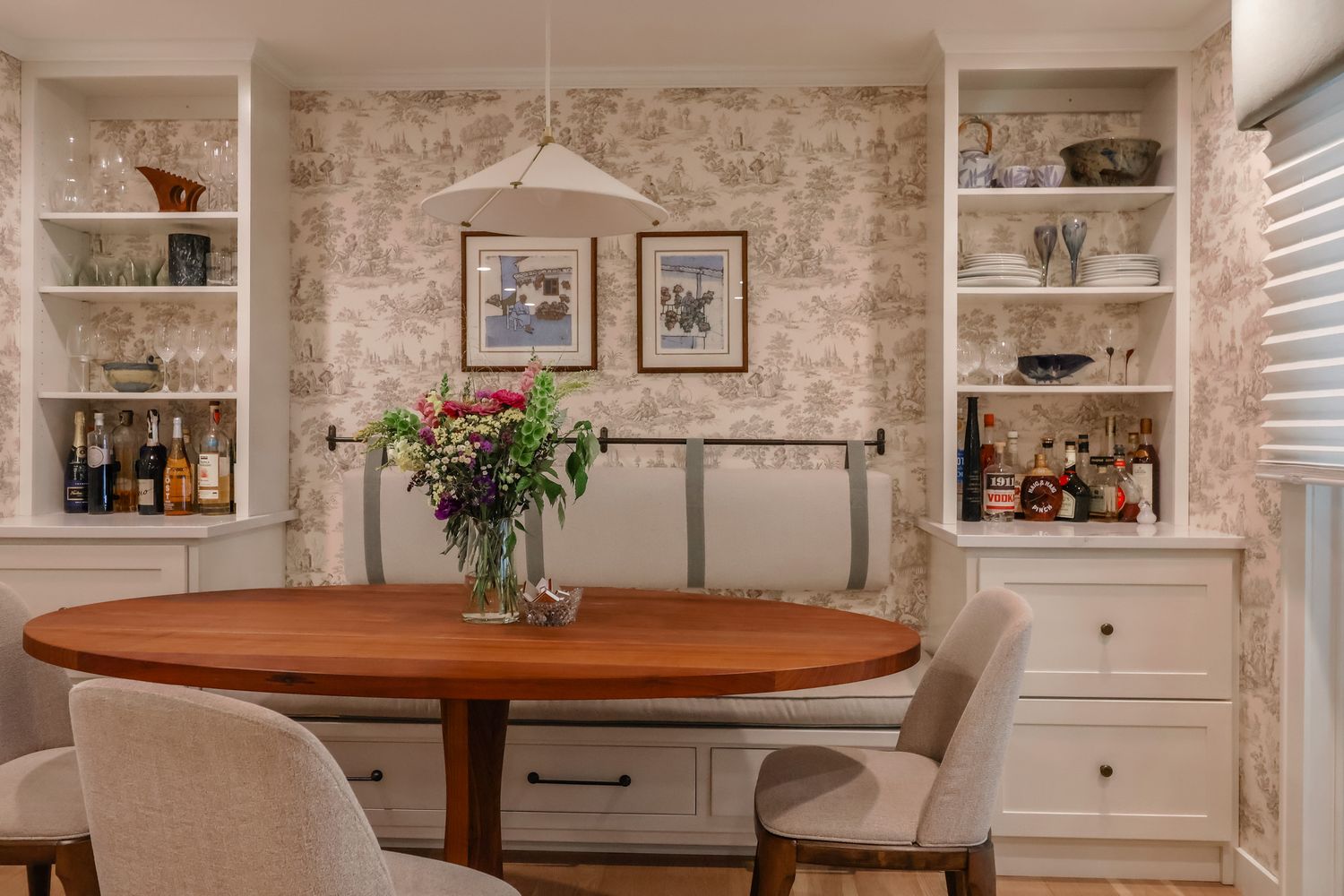 Elegant dining nook with oval wood table, upholstered chairs, built-in shelves, and floral toile wallpaper.