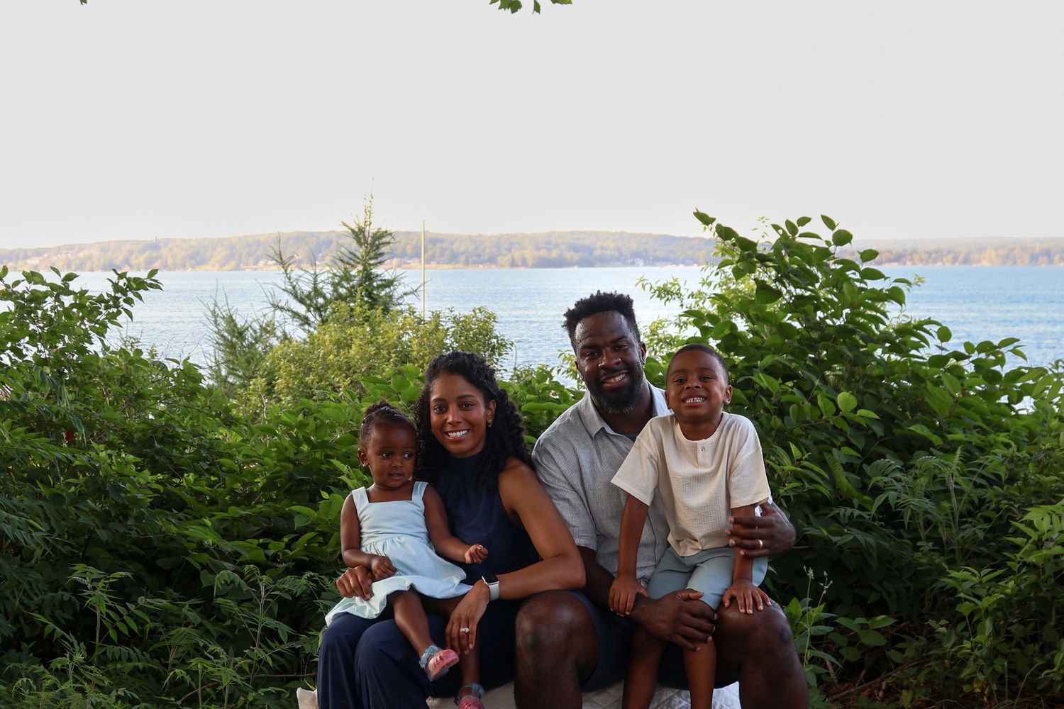 Happy family of four smiling together seated among lush greenery with a scenic waterfront backdrop.