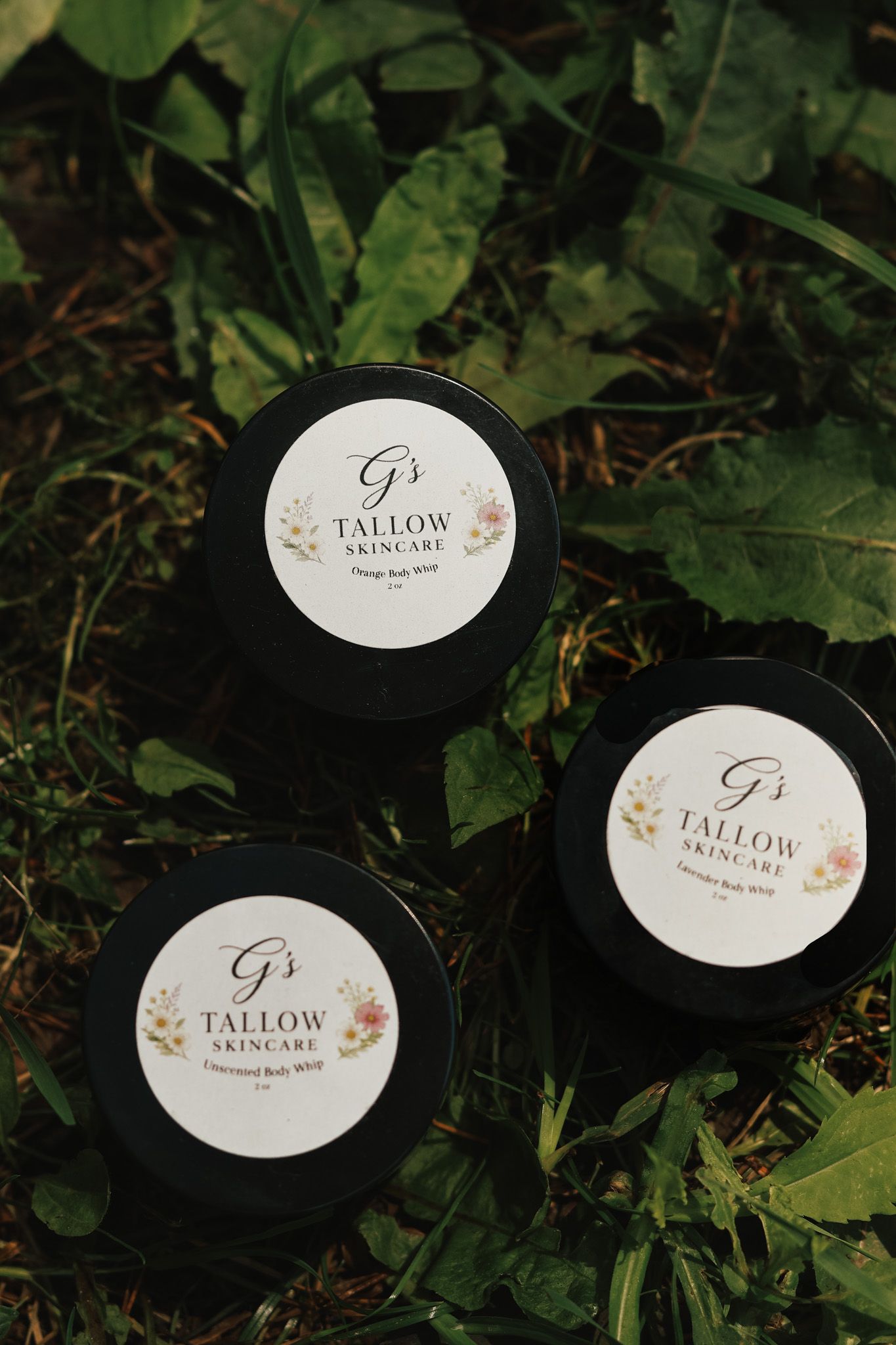 Three black jars of G's Tallow Skincare body whip arranged on green leaves outdoors.