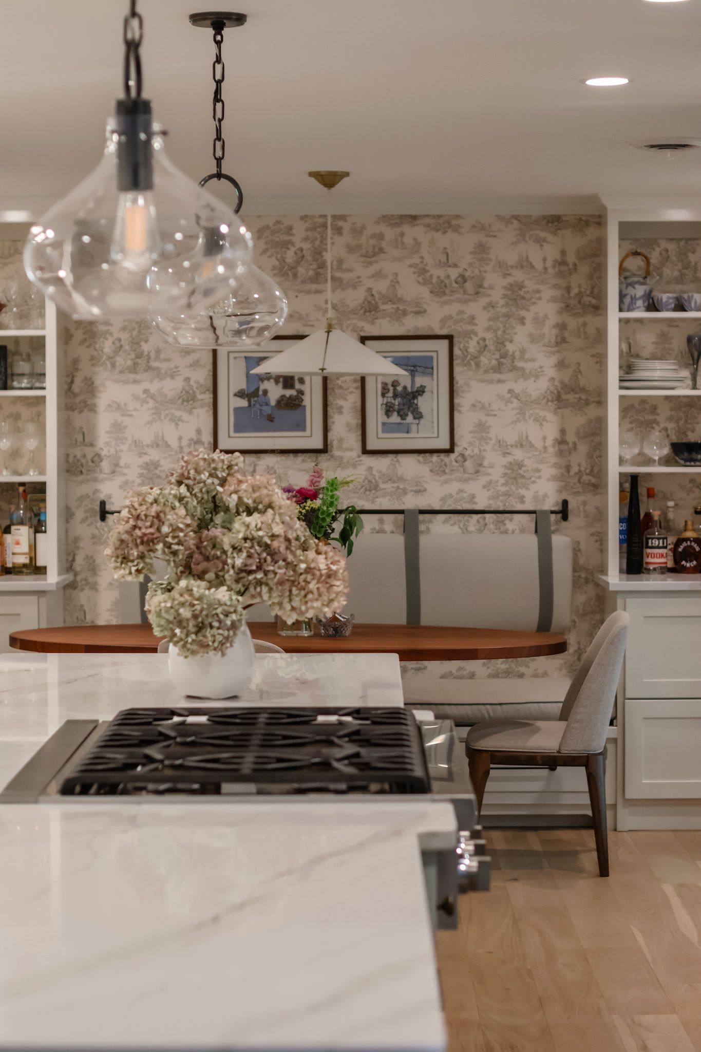 Kitchen island with gas cooktop, glass pendant lights, and cozy banquette dining nook with toile wallpaper.