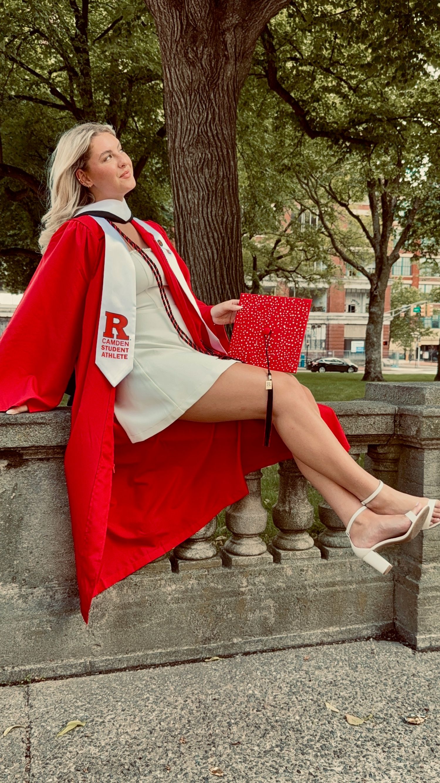 Blonde woman in red Rutgers graduation gown sitting on stone railing under a tree outdoors.